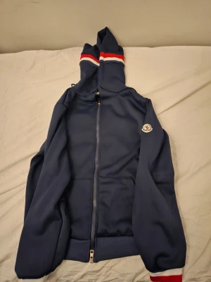Marinblå Moncler hoodie med dragkedja - Snygg marinblå hoodie från Moncler med hel dragkedja framtill och huva. Huven har röda och vita ränder och ärmarna har ribbade muddar. Moncler-logga på vänster ärm. Perfekt för en sportig och stilren look. Helt oanvänd INTE ÄKTA!