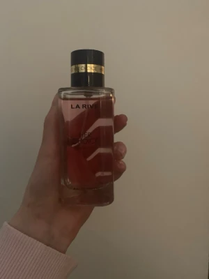 La Rive Her Choice parfym - La Rive Her Choice parfym i en genomskinlig glasflaska med svart och guld kork. Flaskan har en stilren design med rosa toner i vätskan och grafiska vita linjer på framsidan. Perfekt för dig som gillar eleganta och moderna dofter.Helt oanvänd .orgenariepris:300kr