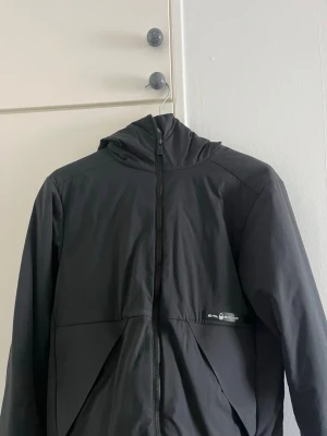  Sail Racing jacka - Snygg svart vindjacka från Sail Racing med huva och hel dragkedja framtill. Jackan har diskret logga på bröstet och en unik detalj på huvan. Tillverkad i lätt, vindtätt material som passar perfekt för blåsiga dagar.