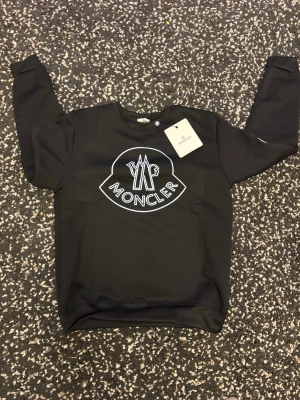 Svart Moncler sweatshirt - Svart sweatshirt från Moncler med stort vitt logotryck på bröstet och en liten Moncler-patch på ärmen. Tröjan har rund halsringning och ribbade muddar vid ärmslut och nederkant. Perfekt för en stilren och trendig look. Den är liten i storlek så den kommer att passa storlek M