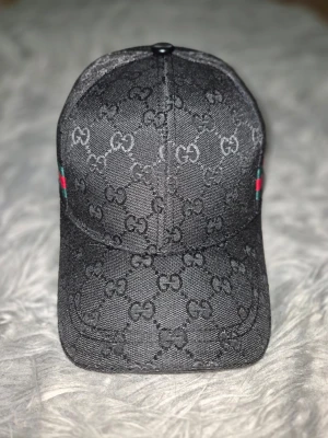 Svart keps från Gucci med GG-mönster - Snygg svart keps från Gucci med det klassiska GG-monogrammet över hela kepsen. På sidorna finns de ikoniska gröna och röda ränderna. Kepsen har böjd skärm och är tillverkad i ett slitstarkt tygmaterial som ger en exklusiv känsla.