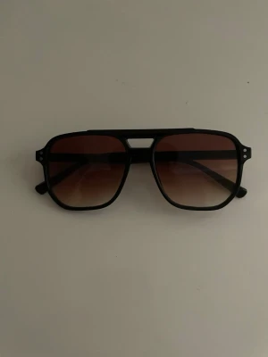 Svarta retro solglasögon - Snygga solglasögon med svart ram och bruna tonade glas. Modellen har en klassisk retroform med dubbla näsbryggor och breda skalmar. Perfekt för dig som vill ha en cool och tidlös look i sommar.