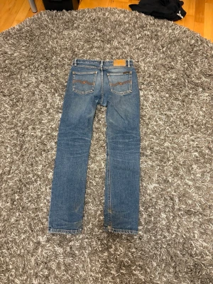 Blå jeans från Nudie Jeans - Säljer ett par klassiska blå jeans från Nudie Jeans med smal passform och fem fickor. Jeansen har snygga slitningar och kontrastsömmar samt läderpatch bak i midjan. 