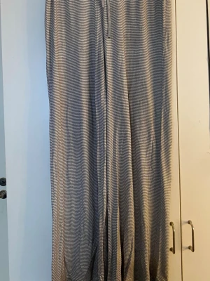 Randiga grå mjukisbyxor med dragsko - Säljer ett par mjukisbyxor med smala svarta ränder på grå bakgrund. Byxorna har en lös passform och dragsko i midjan för extra komfort. Perfekta för chill dagar och avslappnade stunder. Materialet är mjukt och stretchigt.