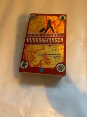 Hundraåringen av Jonas Jonasson - Rolig och äventyrlig roman, 'Hundraåringen som klev ut genom fönstret och försvann' av Jonas Jonasson. Pocketbok med röd och gul omslagsdesign, karta och illustrationer på framsidan. Perfekt för dig som gillar humor och oväntade berättelser.