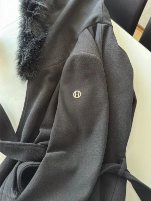 Svart kappa med fuskpäls från Hollies - Säljer denna fina kappa från Hollies. Den är endast använd ett par gånger och i mycket bra skick, men kommer tyvärr inte till användning längre då jag har för många jackor.  Nypris var cirka 9 000 kr, säljs nu för 6 000 kr. Pris kan diskuteras.  Kappan har pälsdetaljer både på luvan och ärmarna, som enkelt går att ta av om man önskar en mer stilren look. Den har även ett knytskärp i midjan så du kan justera passformen efter egen stil.  Perfekt för både höst och vinter. 