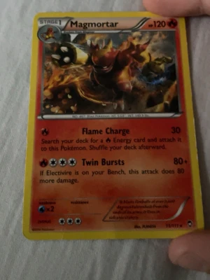 Pokémonkort Magmortar 37/111 - Säljer ett Magmortar Pokémonkort, nummer 37/111. Kortet har en eldig design med Magmortar i centrum och röda, orange och gula färger. Text och effekter på engelska. Baksidan har klassiskt blått Pokémon-motiv med Pokéboll.