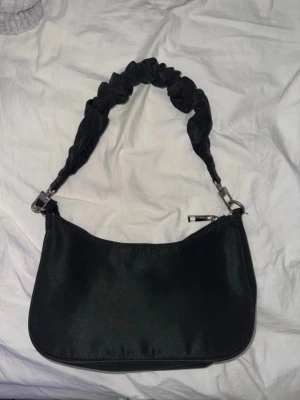 JSvart handväska från BIK BOK - Använt 1 gång. Trendig svart handväska i slät textil med scrunchie-inspirerat axelband. Väskan har en mjukt rundad form och dragkedja upptill. Perfekt för dig som gillar en stilren men ändå lekfull look. Passar till många olika outfits och är lätt att bära.