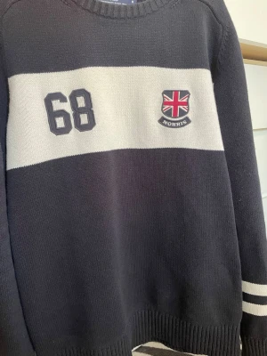 Mörkblå stickad tröja - Snygg mörkblå stickad tröja med bred vit rand över bröstet, broderad siffra och brittisk flagga med logga. Tröjan har rund hals och ribbade muddar. Perfekt för en sportig och stilren look.