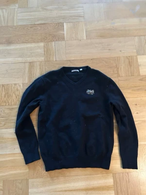 Svart v-ringad tröja Hills Golf Club - Svart v-ringad tröja från Ashworth med Hills Golf Club-brodyr på bröstet. Klassisk passform med långa ärmar och ribbade muddar. Perfekt för dig som gillar stilren och sportig look. Materialet känns mjukt och bekvämt.