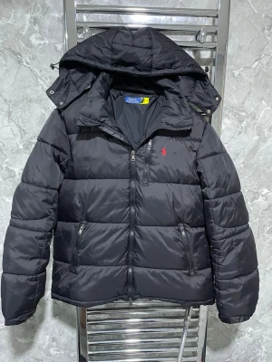 Svart pufferjacka från Polo Ralph Lauren - Snygg svart pufferjacka från Polo Ralph Lauren med avtagbar huva och dragkedja framtill. Jackan har två sidofickor med dragkedja och den klassiska röda loggan broderad på bröstet. Perfekt för kalla vinterdagar och riktigt skön att bära.