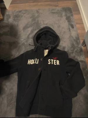 Svart hoodie från Hollister(sällsynt) - Mysig svart hoodie från Hollister med stor huva och knappar framtill. Stora vita bokstäver med Hollister-logga på bröstet och två fickor. Perfekt för en avslappnad och cool look. Tillverkad i mjukt material som känns skönt mot huden.