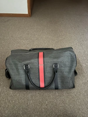 Valentino weekendbag med röd rand - Snygg weekendbag från Valentino i mörkgrått med logotypmönster över hela väskan och en röd vertikal rand i mitten. Väskan har svarta handtag, justerbar axelrem och dragkedja. Perfekt för resor när du vill ha stil och funktion. Tillverkad i slitstarkt syntetmaterial.