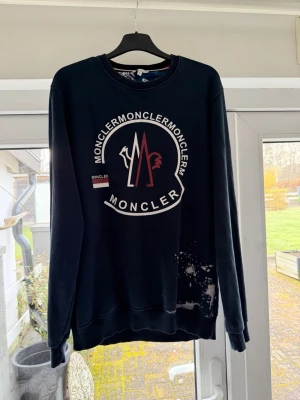 Svart sweatshirt från Moncler - Cool marinblå sweatshirt från Moncler med stort tryckt logomärke på bröstet i vitt och rött. Rund halsringning och ribbade muddar vid ärmslut och nederkant. Tillverkad i mjukt bomullsmaterial, perfekt för en avslappnad streetwear-look.