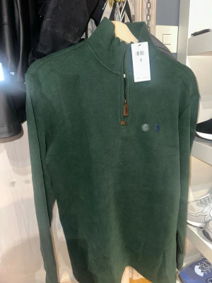 Grön halvzip tröja från Polo Ralph Lauren - Mörkgrön ribbad tröja från Polo Ralph Lauren med halv dragkedja och hög krage. Klassisk blå logga broderad på bröstet. Perfekt för lager på lager och en stilren look. Materialet känns mjukt och bekvämt.