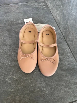 Ljusa rosa ballerinaskor från H&M - Säljer ett par ljusrosa ballerinaskor från H&M med rund tå, dekorativ rosett framtill och ett elastiskt band över foten för extra komfort. Skorna har en klassisk och stilren design i ett mjukt material som ger en söt och trendig look.