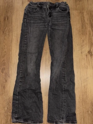Svarta bootcut jeans med slitningar - Ett par svarta lågmidjade bootcut jeans med lätt slitna detaljer och klassisk femficksmodell. Jeansen har en något utsvängd passform nedtill och synliga sömmar. Perfekta för en avslappnad och trendig look.