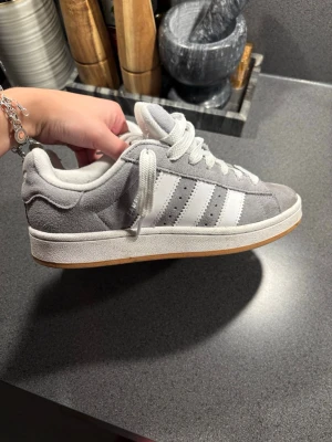 Grå Adidas Campus sneakers - Säljer ett par grå Adidas Campus sneakers i mocka med klassiska vita ränder på sidorna och vit sula med gummidetalj. Skorna har lågt skaft och vita skosnören. Perfekta för en clean och stilren look. Storlek 39