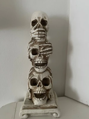 Dekorativ dödskallestatyett - Cool statyett med tre staplade dödskallar där händer täcker ögon, öron och mun. Perfekt som accessoar till maskerad eller cosplay. Tillverkad i hård plast med antikvit finish och detaljerad design.