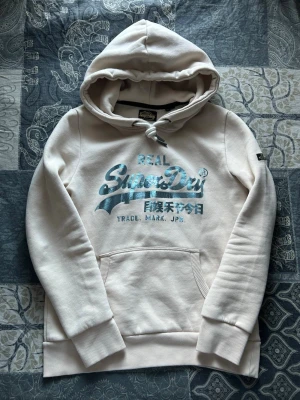 Beige hoodie från Superdry med tryck - Snygg beige hoodie från Superdry med silverfärgat tryck på bröstet och japanska tecken. Klassisk känguruficka framtill, ribbade muddar och dragsko i huvan. Mjuk bomullsblandning som känns skön mot huden. Perfekt för en avslappnad streetwear-look. Skick 10/10, nypris 800