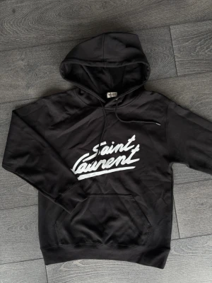 Svart Saint Laurent hoodie - Svart hoodie från Saint Laurent med vit logga tryckt framtill. Klassisk modell med huva, dragsko och magficka. Mjuk bomullskänsla och avslappnad passform, perfekt för en cool streetstyle-look.