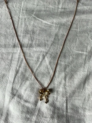 Guldigt halsband med rosettberlock - Snyggt halsband i guldfärgad metall med en söt rosett som hänge. Kedjan är tunn och smidig, och rosetten har en glansig finish som ger ett elegant intryck. Perfekt accessoar för att lyfta din outfit med en feminin touch.