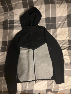Nike Tech Fleece hoodie svart/grå - Nike Tech Fleece hoodie med dragkedja i svart och ljusgrått. Snygg tvåfärgad design, huva och fickor med dragkedja på ärmen. Perfekt för en sportig och stilren look. Materialet är mjukt och stretchigt för extra komfort. Köpt i november, aldrig använd.