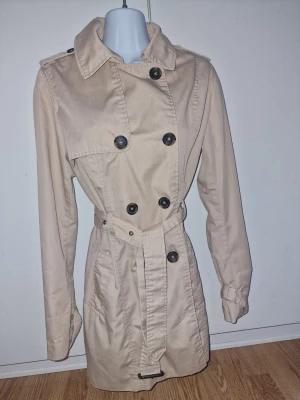 Beige trenchcoat med bälte - Stilren beige trenchcoat med dubbelknäppning och klassisk krage. Tillverkad i ett lätt tyg, perfekt för lager på lager.