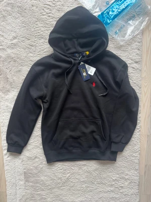 Polo Ralph Lauren hoodie -  svart hoodie från Polo Ralph Lauren med klassisk huva och känguruficka. Ikoniska röda loggan broderad på bröstet. Mjuk bomullsmix och ribbade muddar vid ärmar och nederkant. Perfekt för en avslappnad och stilren look.