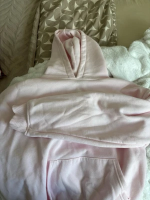 Ljusrosa hoodie med huva - En stilren ljusrosa hoodie med klassisk huva och magficka. Hoodien har ribbade muddar vid ärmsluten och är gjord i mjukt bomullsmaterial. Perfekt för en avslappnad och trendig look.