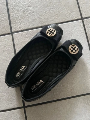 Svarta ballerinaskor med rosett - Snygga svarta ballerinaskor från NE & NA Collection med quiltad innersula, lackdetaljer och dekorativ rosett framtill. På rosetten sitter ett guldfärgat emblem med glittrande stenar som ger skorna en elegant touch. Perfekta för dig som gillar stilrena och trendiga skor.