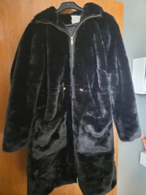 Svart fuskpälsjacka med huva - Snygg svart fuskpälsjacka med huva och dragkedja framtill. Jackan har två stora fickor och detaljer i guld, vilket ger en lyxig känsla. Perfekt för kalla dagar när du vill hålla dig varm och samtidigt se trendig ut. NY utan lappen kvar