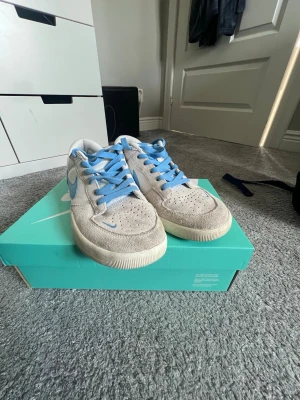 Nike SB Dunk Low grå/blå sneakers - Nike SB Dunk Low sneakers i grå mocka med ljusblå detaljer och snören. Skorna har perforerad tåbox för ventilation och klassisk Nike Swoosh på sidan. Yttersulan är vit och skon har en låg silhuett som passar streetwear-stilen.