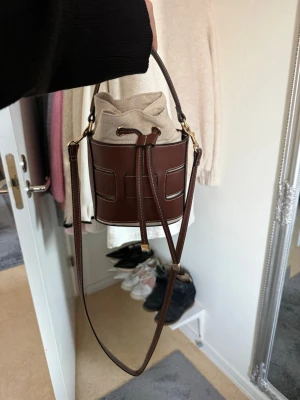 Brun bucket bag med canvasdetaljer - Säljer en snygg brun bucket bag med vita sömmar och justerbar axelrem. Väskan har en unik cylinderform och stängs med en beige canvaspåse med dragsko. Perfekt för dig som gillar trendiga accessoarer med cool design.