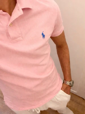 Rosa Piké från Ralph Lauren (Slim Fit) - Säljer en ljusrosa pikétröja från Polo Ralph Lauren med klassisk krage, två knappar vid halsen och blå logga på bröstet. Tröjan är kortärmad och gjord i mjuk bomull med normal passform. Perfekt för en clean och stilren look. Storlek Small slim fit. Snabb Leverans!