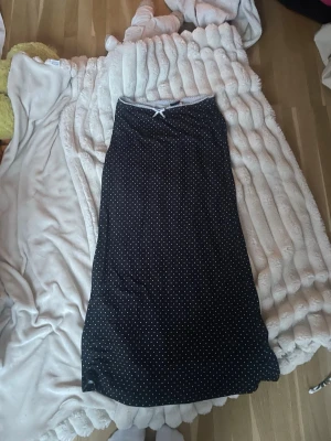 Svart prickigt nattlinne med rosett - Svart långkjol med små vita prickar och en söt liten rosett vid halsringningen. Kjolen har en enkel, rak/ tajt passform Perfekt för dig som gillar klassiska mönster med en twist.