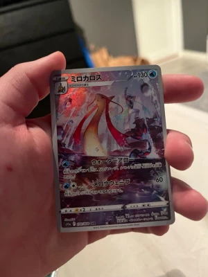 Japanskt Pokémonkort Milotic - Säljer ett japanskt Pokémonkort med Milotic och en tränare på framsidan. Kortet har en holografisk yta med färger i blått, rött och lila. Texten är på japanska och kortet har vattenenergi-symbol. Perfekt för samlare av Pokémonkort.