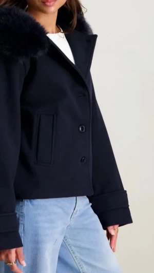Marinblå kort rock med fuskpäls - Snygg marinblå kort rock med stora slag och fuskpälsdetalj på kragen. Rocken har dubbelknäppning framtill, raka fickor och breda ärmslut. Perfekt för dig som vill ha en stilren och trendig look i höst och vinter.