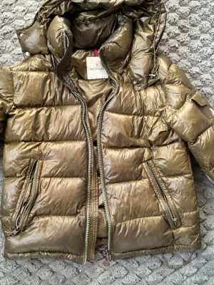 Olivgrön dunjacka från Moncler - Säljer en olivgrön dunjacka från Moncler med glansig finish och huva. Jackan har två snedställda fickor med dragkedja, hög krage och quiltad design. Perfekt för kalla vinterdagar och riktigt snygg till streetwear-stilen. STORLEK 1/s MEN PASSAR ÄVEN M