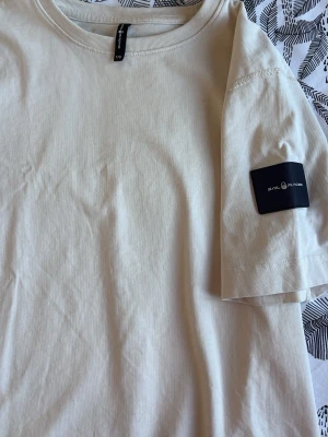 Beige t-shirt från Sail Racing - Snygg beige t-shirt från Sail Racing med logga på ärmen och diskret tryck på sidan. Klassisk rund hals och korta ärmar. Perfekt för en clean och stilren look.