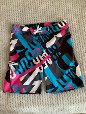 Färgglada boardshorts med tryck - Säljer ett par snygga boardshorts med grafiskt mönster i svart, turkos, rosa och vitt. Shortsen har snörning i midjan och en sidoficka med dragkedja. Perfekta för stranden eller poolen. Materialet känns som snabbtorkande polyester.