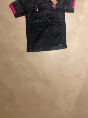 Svart Nike fotbollströja Palmer 20 - Säljer en svart fotbollströja från Nike med rosa detaljer på ärmarna och gula inslag vid sidorna. Tröjan har trycket 'PALMER 20' på ryggen och Premier League-logga. Perfekt för dig som älskar fotboll och vill sticka ut på planen.