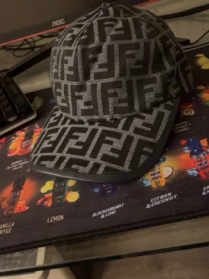 Svart keps med Fendi-logga - Säljer en svart keps från Fendi med det ikoniska FF-mönstret i grått över hela kepsen. Klassisk böjd skärm och justerbar baksida. Tillverkad i ett slitstarkt tygmaterial som ger en exklusiv känsla och streetwear-vibe. Jag säljer den eftersom den tyvärr inte passar min huvudform och lite för stor för mig. Använt den 2 gånger