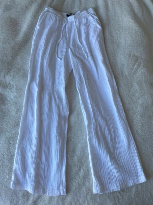 Vita vida linnebyxor - Säljer ett par vita linnebyxor med avslappnad passform och dragsko i midjan. Byxorna har raka, vida ben och är tillverkade i ett luftigt, strukturerat tyg som känns perfekt för varma dagar. Superenkla att styla och riktigt sköna att bära.