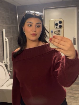 Vinröd off shoulder stickad tröja - Mysig vinröd stickad tröja med off shoulder-design och bred ribbad kant vid halsen. Tröjan har långa ärmar och en avslappnad passform, perfekt för en trendig och bekväm look. Materialet ser mjukt och fluffigt ut.