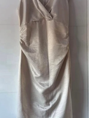 Ny.Champagnefärgad omlottklänning Vero Moda - Snygg champagnefärgad klänning från Ellos med omlottdetalj och lätt glansigt tyg. Klänningen har korta ärmar, v-ringning och en smickrande drapering framtill. Perfekt för dig som vill ha en elegant look med modern touch. Längd 105cm.