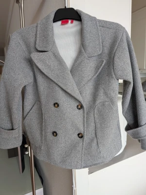 Grå jacka från RYVLS - Stilren ljusgrå vårjacka från RYVLS, modell "Stockholm Peacoat" köpt på Kids Brand Store med dubbelknäppning och stora slag. Jackan har två framfickor och en rem vid ärmslut. Tillverkad i ett mjukt, ull-liknande material som ger en klassisk look. Jättefint skick. Stlk 146/152 men bör även passa XXS. (Ev även XS men kan bli kort i ärmarna då) Skickar snabbt