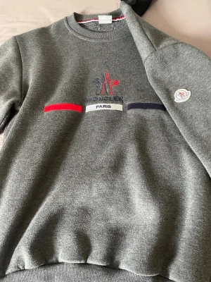 Grå sweatshirt från Moncler Paris - Säljer en grå sweatshirt från Moncler Paris med broderad logga och text på bröstet i rött, blått och vitt. Tröjan har rund halsringning, ribbade muddar och en patch på ärmen. Perfekt för dig som gillar stilrena och exklusiva plagg. Nypris ungefär 2000 men jag är inte säker, priset kan diskuteras 🙂