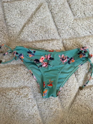 Blommiga bikiniunderdel från Pieces - Mintgrön bikiniunderdel från Pieces med blommigt mönster i rosa, orange och mörkblå toner. Modellen har knyt i sidorna för justerbar passform och är tillverkad i mjuk polyester. Perfekt för strandhäng eller poolparty!