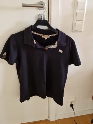 Mörkblå Burberry pikétröja - Snygg svart pikétröja från Burberry med klassiskt rutigt mönster på insidan av kragen och ärmslut. Tröjan har korta ärmar, liten broderad logga på bröstet och är gjord i mjuk bomull. Perfekt för en stilren och avslappnad look. Inge defekter. Inte äkte men av bra kvalité